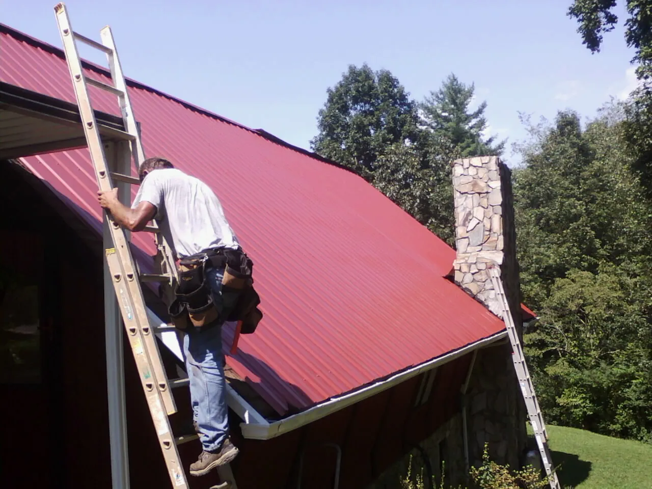 Roofer on a ladder beside a red metal roof and stone chimney — Soffit & Fascia Repair by Roofing Brothers in Oak City